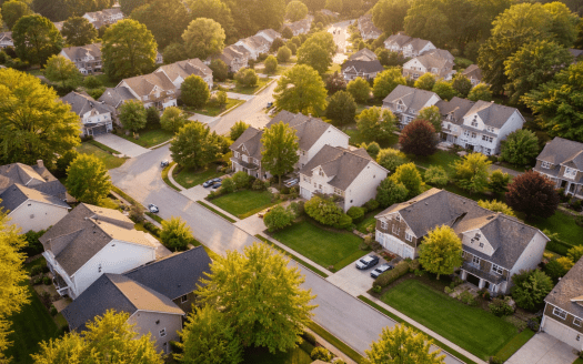Aerial view of a quiet suburban neighborhood with tree-lined streets and well-kept single-family homes, captured in warm golden-hour light.